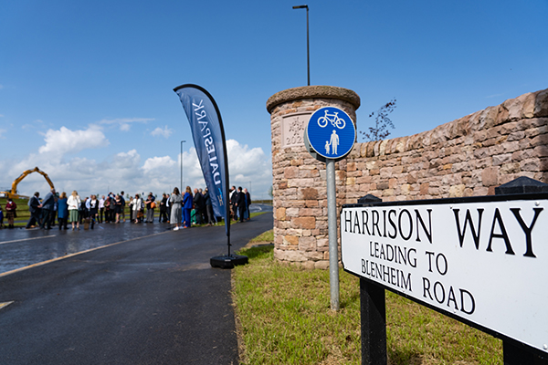 Sign for Harrison Way with the road opening ceremony in the background