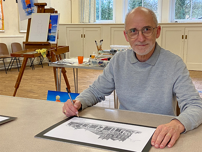 Artist Alan fernihough signs his limited edition print of ashbourne methodist church. Alan is a middle aged white man with a bald head, round glasses and short grey beard.